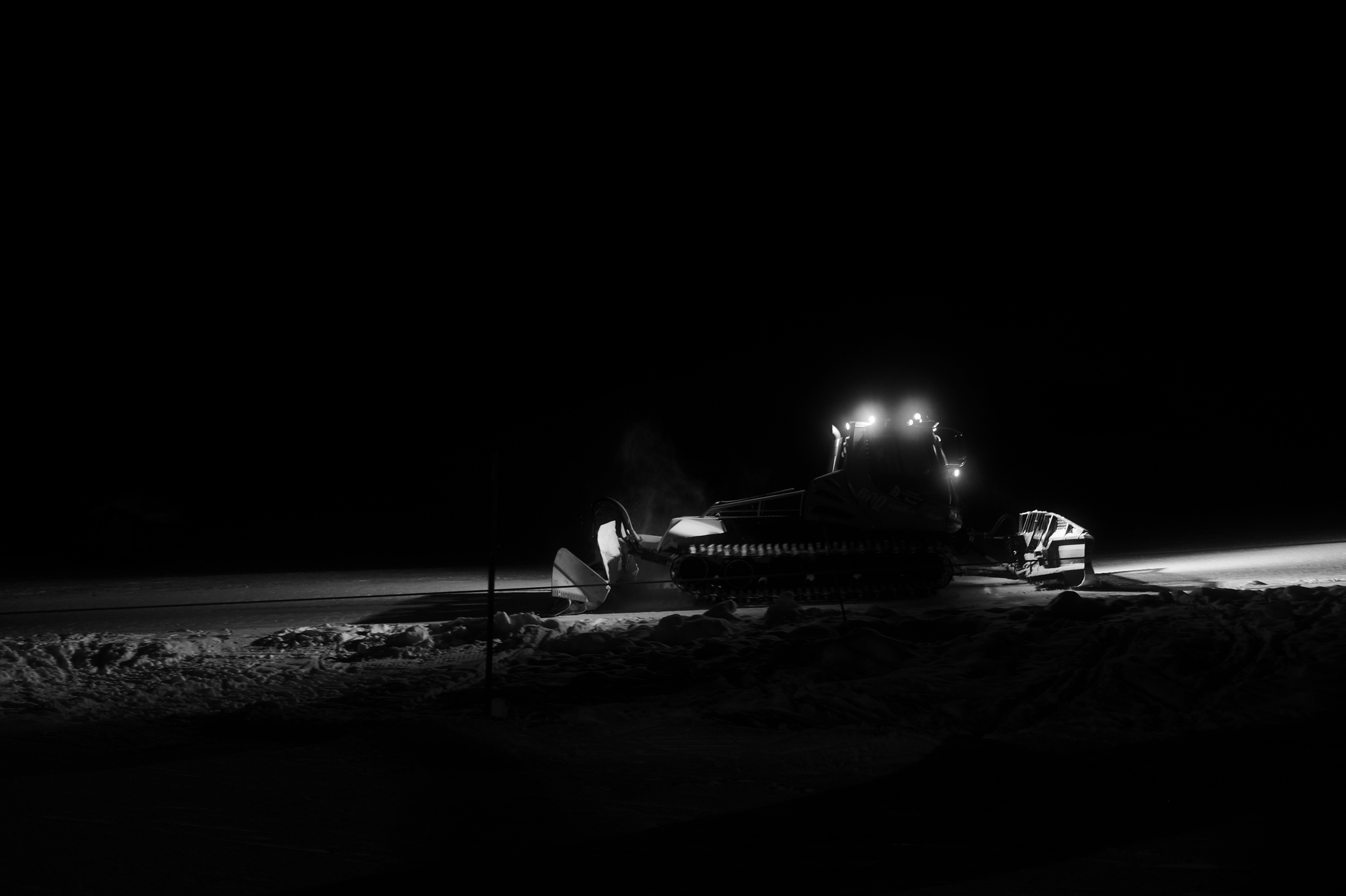 A snow groomer working at night, headlights cutting across groomed snow