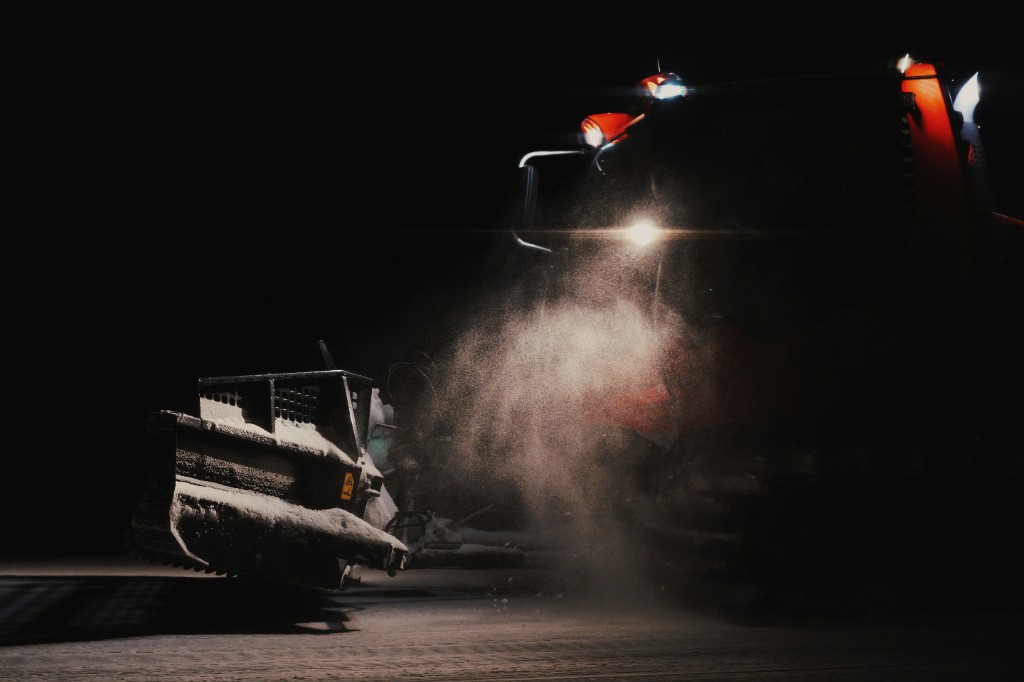 A red snow groomer at night, work lights illuminating snow dust in front of the blade